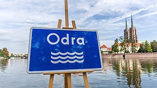 Blaues Schild mit der Aufschrift "Odra" mit Wasser und Stadt im Hintergrund