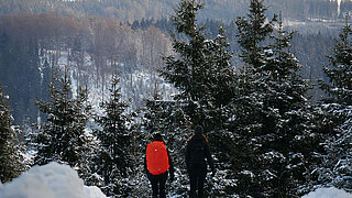 Zwei Personen stehen mit dem Rücken zur Kamera im Schnee; vor ihnen liegt ein verschneites und bewaldetes Tal.