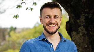 Porträt eines freundlich lächelnden Mannes mit kurzem braunem Haar und Bart vor einem Baum. Er trägt ein blaues Hemd, im Hintergrund sind grüne Wiesen und Bäume in einer leicht verschwommenen, natürlichen Landschaft zu sehen.