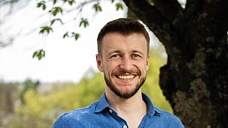 Porträt eines freundlich lächelnden Mannes mit kurzem braunem Haar und Bart vor einem Baum. Er trägt ein blaues Hemd, im Hintergrund sind grüne Wiesen und Bäume in einer leicht verschwommenen, natürlichen Landschaft zu sehen.