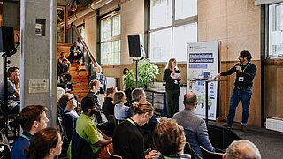 Auf einer Bühne sprechen zwei Personen miteinander, davor sitzen andere Personen, die zuschauen und zuhören. 