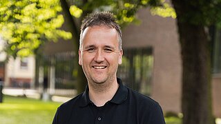 Ein Mann (Fabian Giese) steht vor einem Haus mit Bäumen und lächelt in die Kamera. Er trägt ein schwarzes Shirt. 