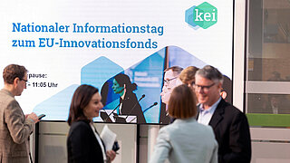 Eine Menschengruppe im Gespräch vor einem großen Bildschirm auf dem geschrieben steht: Nationaler Informationstag zum EU-Innovationsfonds
