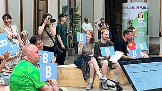 Personen sitzen vor einer Bühne und halten Schilder mit dem Buchstaben B in der Hand