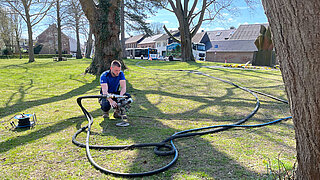 Person kniet auf einer Wiese zwischen Bäumen und hält ein Gerät, an dem mehrere Schläuche angeschlossen sind