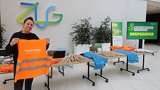 Eine Person steht neben einem Tisch mit Materialien und zeigt eine Warnweste