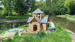 Mann bemalt hölzernes Haus für Wasservögel auf einer grasbewachsenen Insel in einem Teich