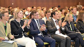 Ein Publikum sitzt auf Stühlen und applaudiert. 