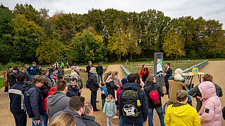 Gruppe von Erwachsenen und Kindern versammelt sich auf einem neu gestalteten, ökologisch aufgewerteten Parkplatz am Spielkaulenweg, umgeben von Bäumen und Grünflächen, mit einer Person, die vor einem Informationsschild spricht.