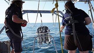  Zwei Personen auf einem Boot bedienen ein hängendes wissenschaftliches Messgerät über dem Wasser.