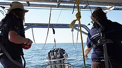  Zwei Personen auf einem Boot bedienen ein hängendes wissenschaftliches Messgerät über dem Wasser.