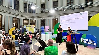 Eine Bühne mit Bildschirm, auf der Personen stehen, davor Publikum