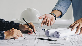 Auf einem Tisch liegt Papier, man sieht Hände, die darauf schreiben oder zeichnen, während die Hände einer anderen Person darauf zeigen