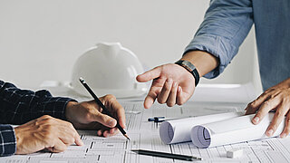 Auf einem Tisch liegt Papier, man sieht Hände, die darauf schreiben oder zeichnen, während die Hände einer anderen Person darauf zeigen