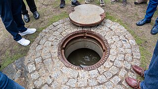 Geöffneter Kontrollschacht der Dükerleitung mit sichtbarem Wasser. Mehrere Teilnehmende des Pressetermins stehen um die Anlage und informieren sich über die Wasserzufuhr für das Moorgebiet.