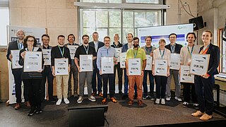 Mehrere Personen stehen auf einer Bühne und zeigen ihre Urkunden in die Kamera