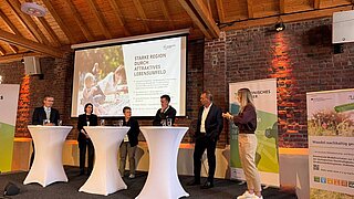 Mehrere Personen stehen an Stehtischen, neben und hinter ihnen Rollups und eine Projektionsfläche