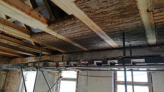 Decke mit freiliegenden Holzbalken und Dämmmaterial, darunter Gerüst und Fenster in einem Rohbau