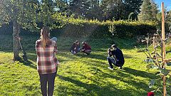 Mehrere Personen stehen bzw. hocken auf einer blühenden Grünfläche, eine Person hält eine Kamera. Im Hintergrund eine Hecke, man sieht ebenfalls einige Obstbäume