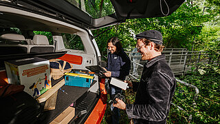 Zwei Männer stehen an der Kofferraumklappe eines Busses. Im Inneren des Fahrzeugs befinden sich Gerätschaften. 