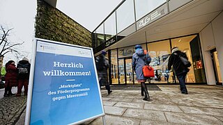 2 Personen gehen in ein Bürogebäude, vor dem Gebäude steht ein Schild, das auf eine Veranstaltung hinweist. 