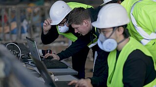 Drei Personen in Schutzkleidung sitzen am Tisch, vor ihnen Laptops, dahinter eine Abfallsortieranlage