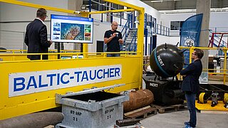 Ein Mann (Carsten Schneider) steht vor einer Art Bühne zum Tauchen. Auf der Bühne befinden sich zwei weitere Personen