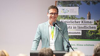 Ein Mann steht an einem Rednerpult und spricht in ein Mikro, hinter ihm ein Poster 