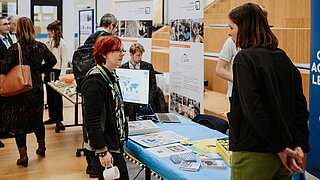 Mehrere Personen stehen an einem Infotisch und schauen sich Dokumente und andere Materialien an, im Hintergrund sieht man weitere Infostände. 