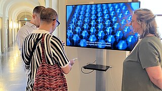 Besucherinnen stehen vor einem großen Bildschirm und sprechen miteinande.r