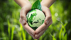 zwei Hände halten eine grüne Weltkugel aus Glas, im Hintergrund Gräser