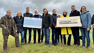 Gruppe von Menschen steht auf einer Wiese, eine Person hält ein großes Schild mit der Aufschrift 'Naturschutzgroßprojekt MITTELELBE SCHWARZE ELSTER', eine andere Person hält einen großen symbolischen Scheck
