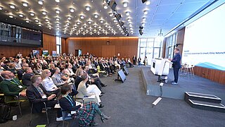 Ein Publikum sitzt vor einer Bühne. Auf der Bühne steht eine Person und spricht, im Hintergrund ein Bildschirm mit einer Präsentation