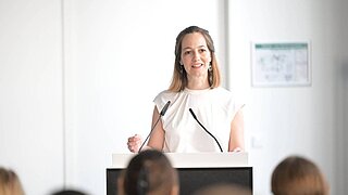 Eine Frau steht an einem Rednerpult. Vor ihr sieht man ein Publikum. 