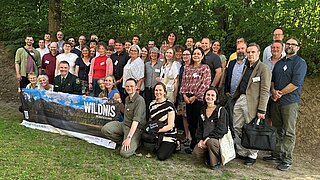 Gruppenbild mit vielen stehenden und knieenden Personen vor Bäumen. Einige halten ein Spruchband in der Hand, auf dem "Wildnis" steht