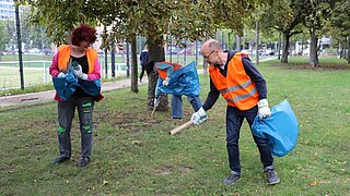 Drei Personen stehen auf einer Grünfläche und sammeln Unrat