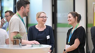 Drei Personen stehen an einem Stehtisch und reden miteinander. Im Raum sind weitere Personen