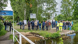 Am Ufer der Havel stehen Teilnehmende der Eröffnungsfeier und blicken in Richtung des Redepults. Am Wasserrand ist der Zufluss zur Dükerleitung durch eine blaue Markierung gekennzeichnet.