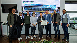 8 Personen stehen auf einer Bühne, eine Person hält eine Urkunde in den Händen. Im Hintergrund sieht man ein großes Foto mit der Überschrift "Dachbegrünung Borussia-Park"