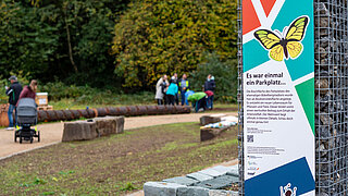 Blick auf eine Grünfläche mit Wegen. Im Vordergrund ein Schild, das erklärt, dass sich dort einmal ein Parkplatz befand