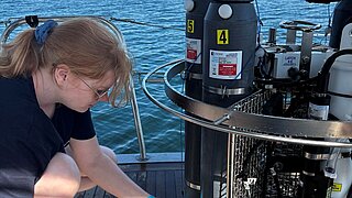 Frau mit blonden Haaren und blauen Handschuhen entnimmt Flüssigkeit aus wissenschaftlichem Gerät auf Boot mit Meer im Hintergrund