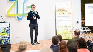 Ein Mann steht auf einer Bühne und spricht, vor ihm sitzen Personen. 