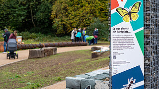 Informationsschild zur ökologischen Aufwertung des ehemaligen Reisebus- und Presseparkplatzes am Bökelbergstadion, neben einem Rosenbeet und einem Weg, im Hintergrund unscharf mehrere Personen und ein Kinderwagen.