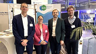 Gruppenbild: die verantwortlichen Projektmitarbeitenden des Servicezentrum stehen neben den KEI Kollegen Max Mileweski und Matthias Heck an einem Stechtisch, im Hintergrund ist ein KEI Roll-up zu sehen.