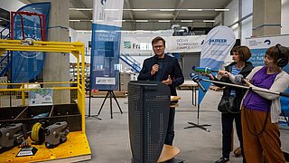 Ein Mann (Carsten Schneider) steht an einem Rednerpult, seitlich von ihm andere Personen mit Mikros. 