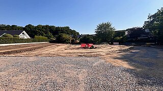 Eine Sand- und Schotterfläche, im Hintergrund Bäume und Gebäude, auf der Fläche Zaunteile zum Absperrender Fläche ein F