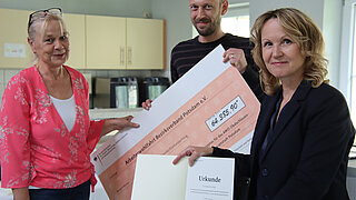 Drei Personen halten einen großen Förderscheck in den Händen und blicken in die Kamera.