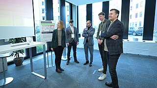 Mehrere Personen stehen vor einem Tisch und einer Leinwand und bekommen etwas vorgeführt