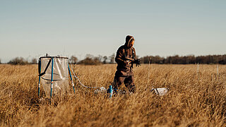 Person steht in einem weiten Feld mit hohem Gras neben einem großen, transparenten Behälter mit blauen Halterungen und hält ein Tablet oder ähnliches Gerät in den Händen