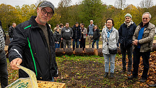 Gruppe von Erwachsenen steht auf einer herbstlichen, ökologisch umgestalteten Grünfläche mit Baumstämmen im Hintergrund.